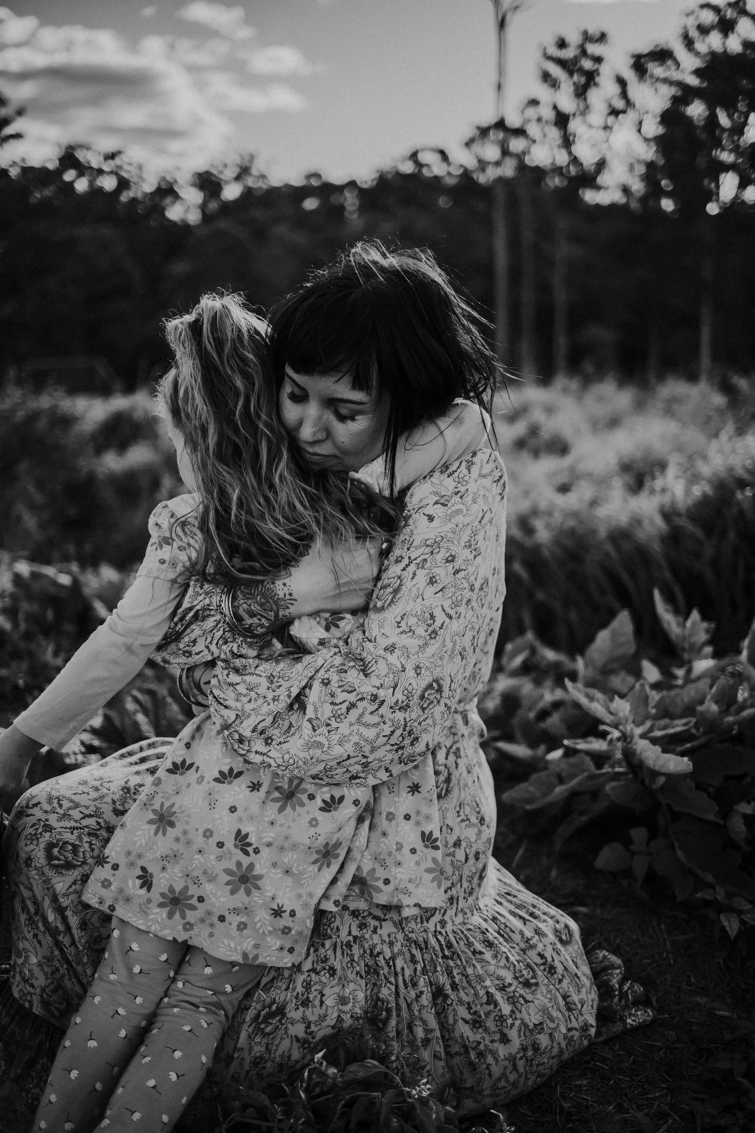 A woman with short dark hair embracing a young girl with long wavy hair outdoors in a field, both wearing patterned dresses.