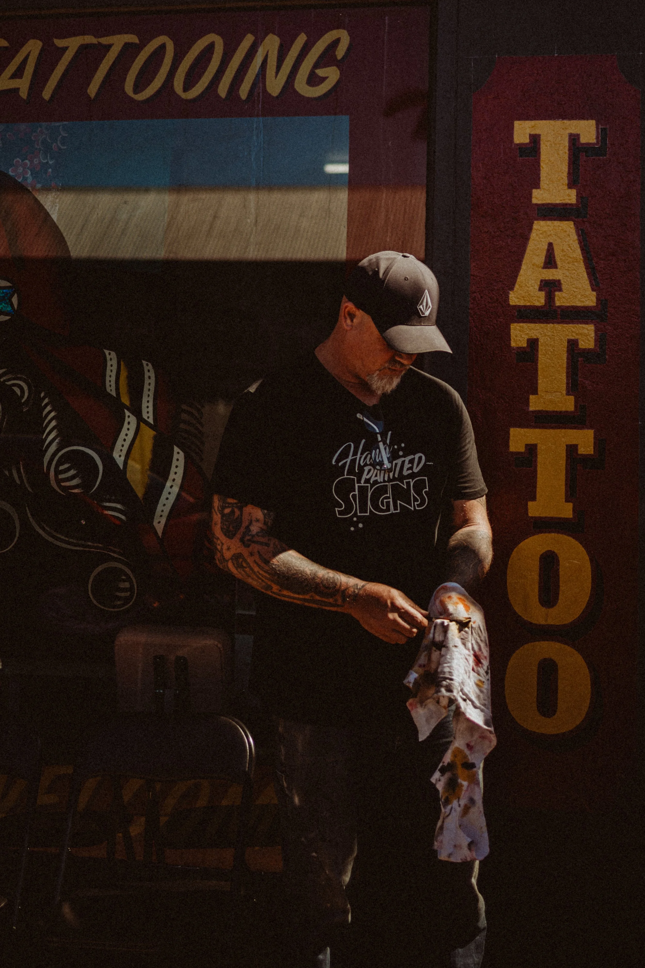 A man with tattoos, wearing a baseball cap and black T-shirt that reads 'Hand Painted Signs', stands under a maroon 'TATTOO' sign, holding a cloth and a tool in his hands.