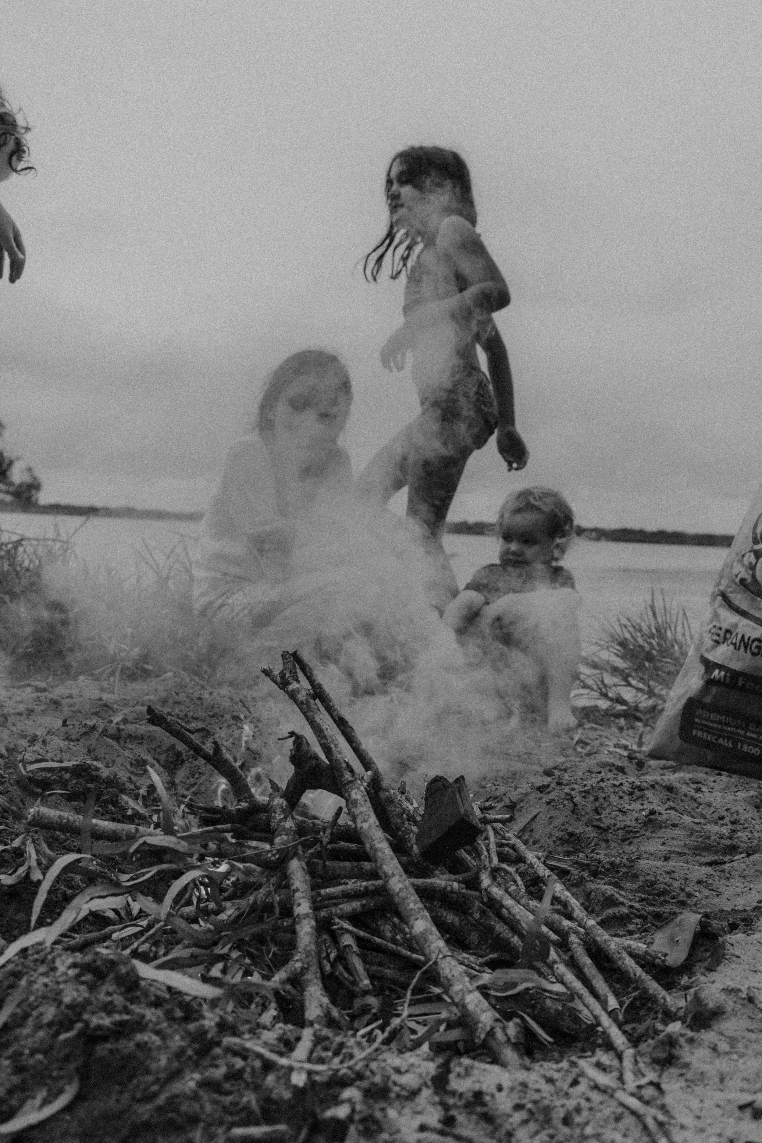 Children playing by a campfire on the beach during daytime, with smoke rising from the fire and a body of water in the background.