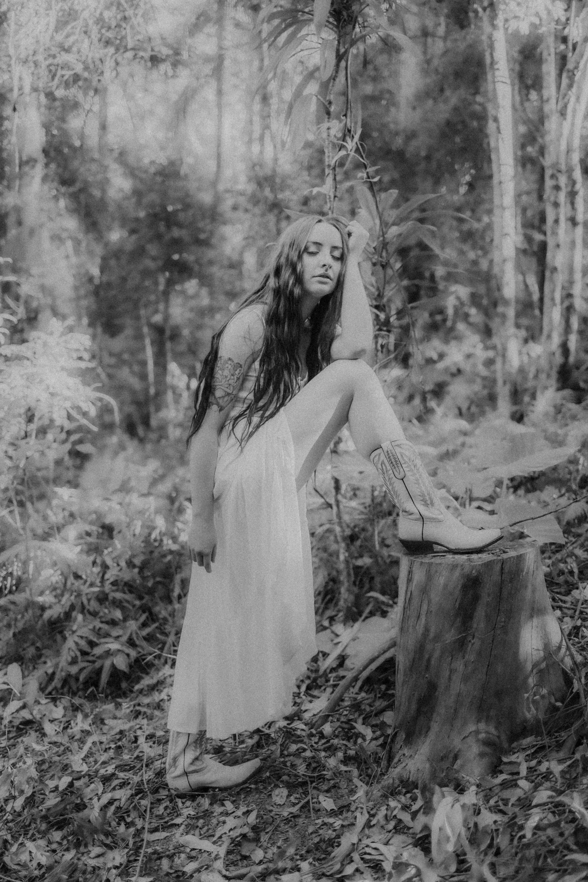 A woman with long dark hair and tattoos on her arm is sitting on a tree stump in a forest. She is wearing a long dress and cowboy boots, with one foot resting on the stump. She has a contemplative expression and is resting her head on her hand, surro
