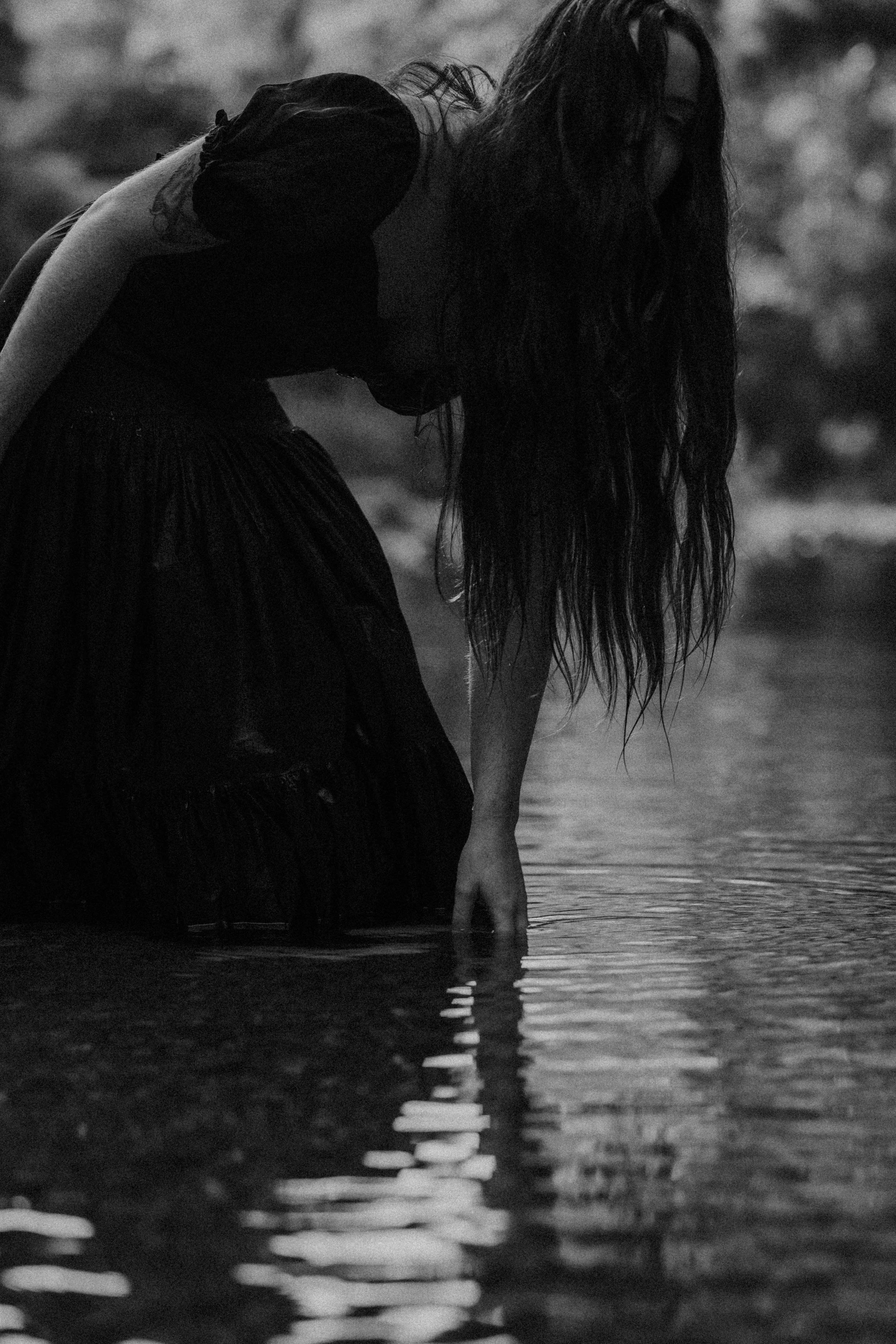 A woman with long hair in a dark dress is kneeling in shallow water, touching the surface with her hand, in black and white.
