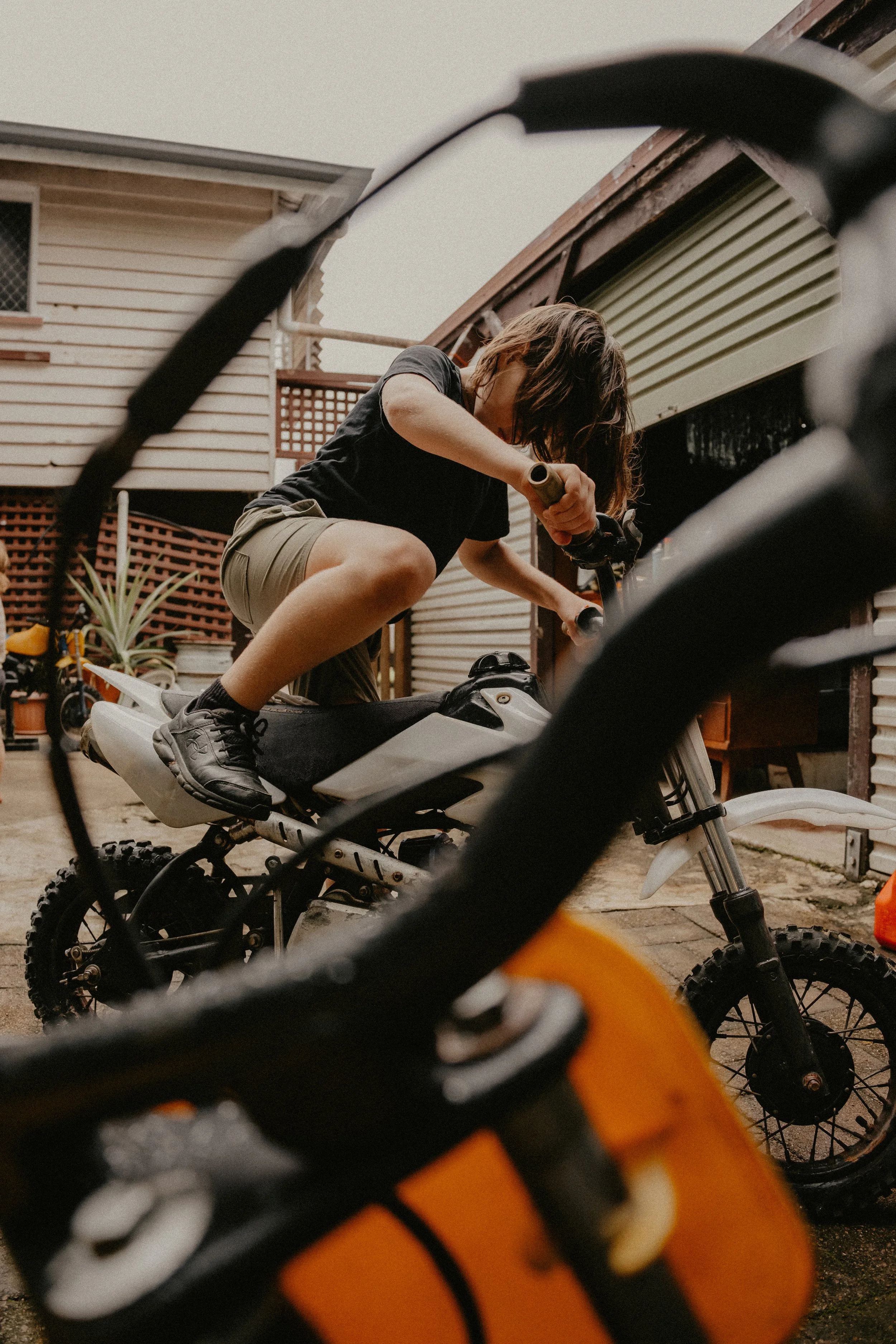 Person with short hair climbing onto a dirt bike in a backyard, with another dirt bike and house in the background, viewed through the handlebars of an orange motorcycle.