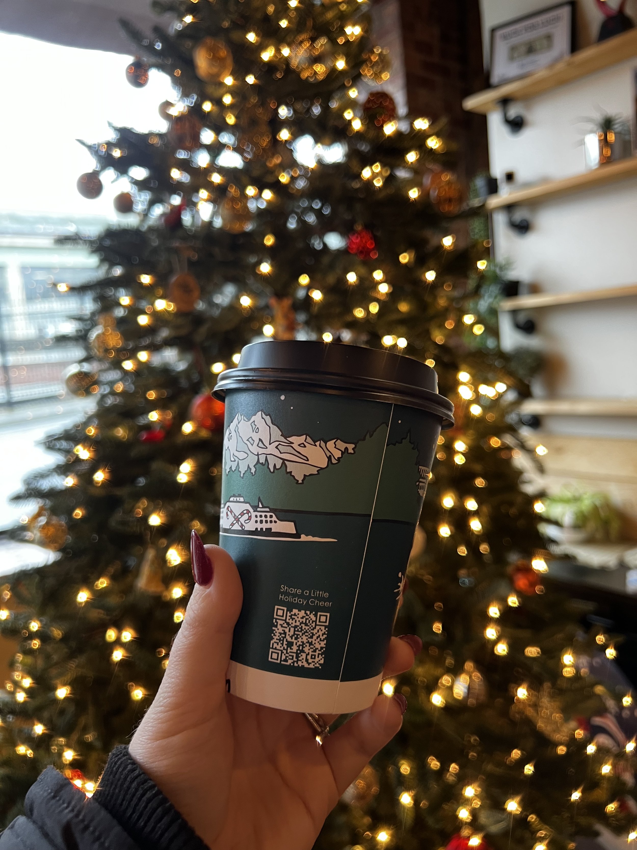 A hand holding a to-go coffee cup in front of a decorated Christmas tree with warm yellow lights.
