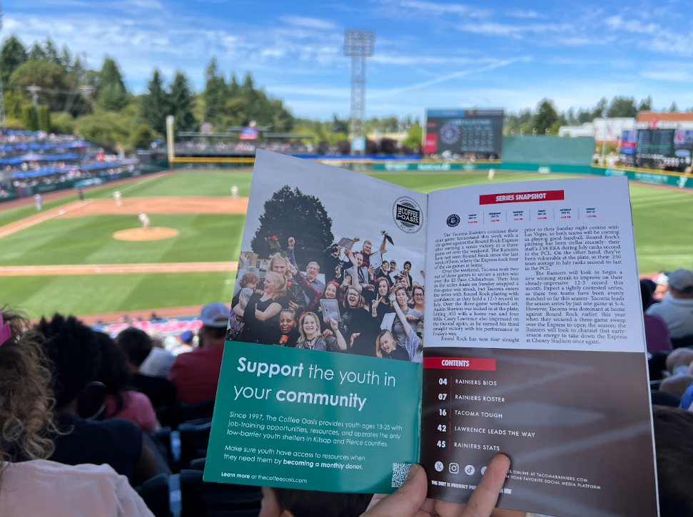 A person holding a community support brochure at a baseball game. The background shows the field with players and a scoreboard on a sunny day.