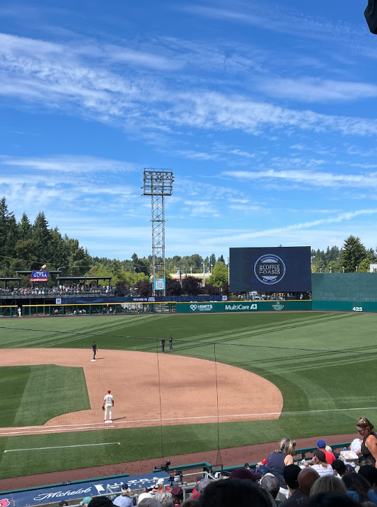 A baseball game is taking place at a stadium with a green field, the infield and outfield visible, and a few players standing on the field. There are spectators in the stands, and a large electronic scoreboard displays advertisements and game information. The sky is clear with some clouds.