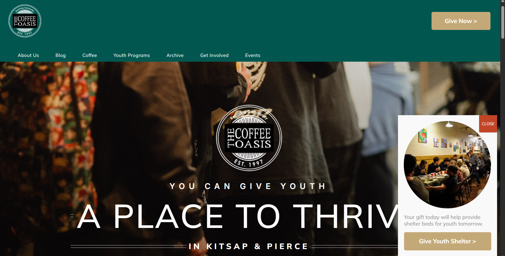 The website header of The Coffee Oasis charity organization featuring its logo, a group of people in a social gathering, and options such as About Us, Blog, Coffee, Youth Programs, Archive, Get Involved, and Events. There is also a prominent Give Youth Shelter button, a circular inset showing a room with people sitting around tables, and overlay text promoting youth giving and support for shelter beds.