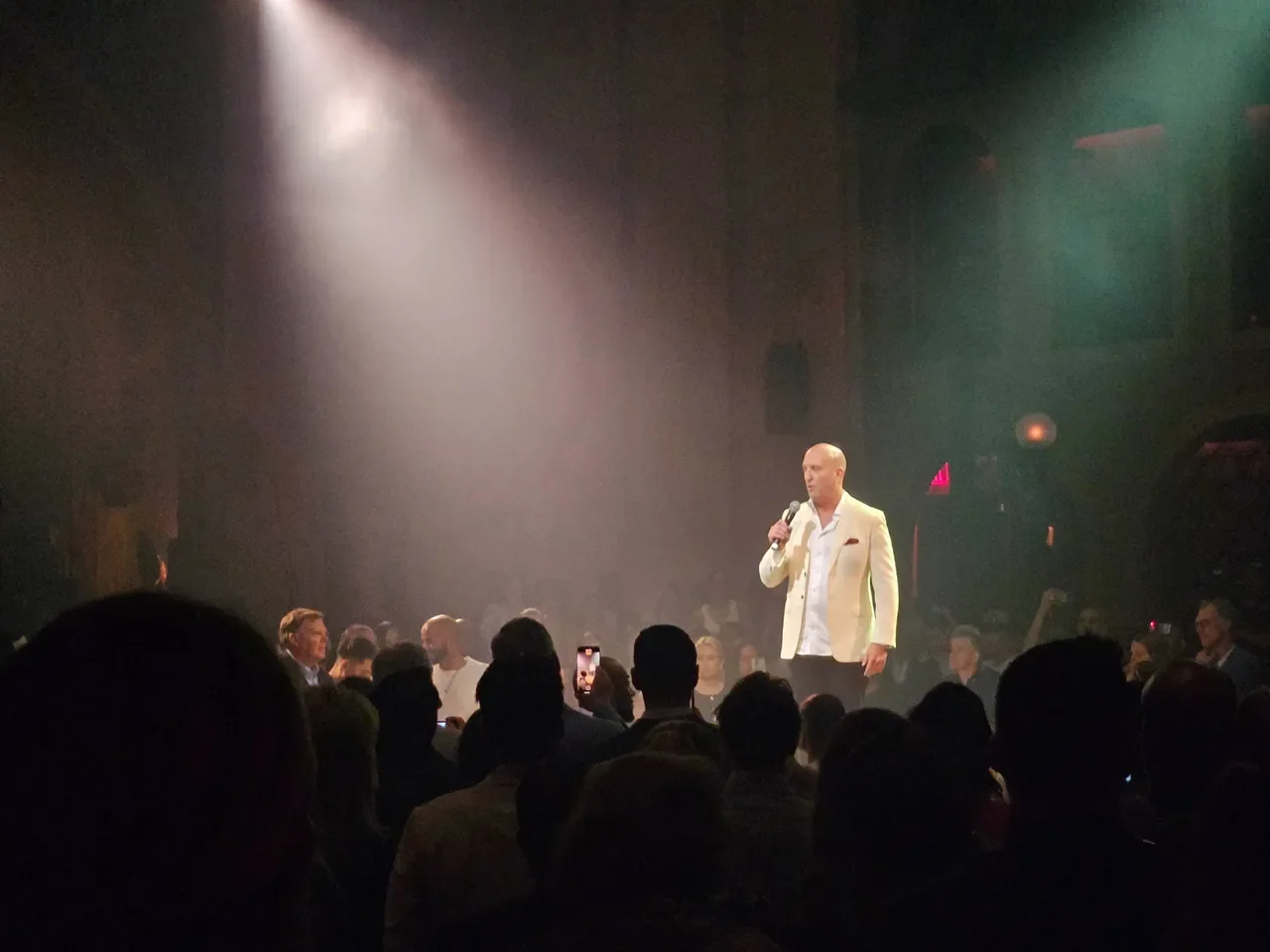 A man in a white blazer speaking into a microphone on a stage with a spotlight, surrounded by an audience in a dark venue.