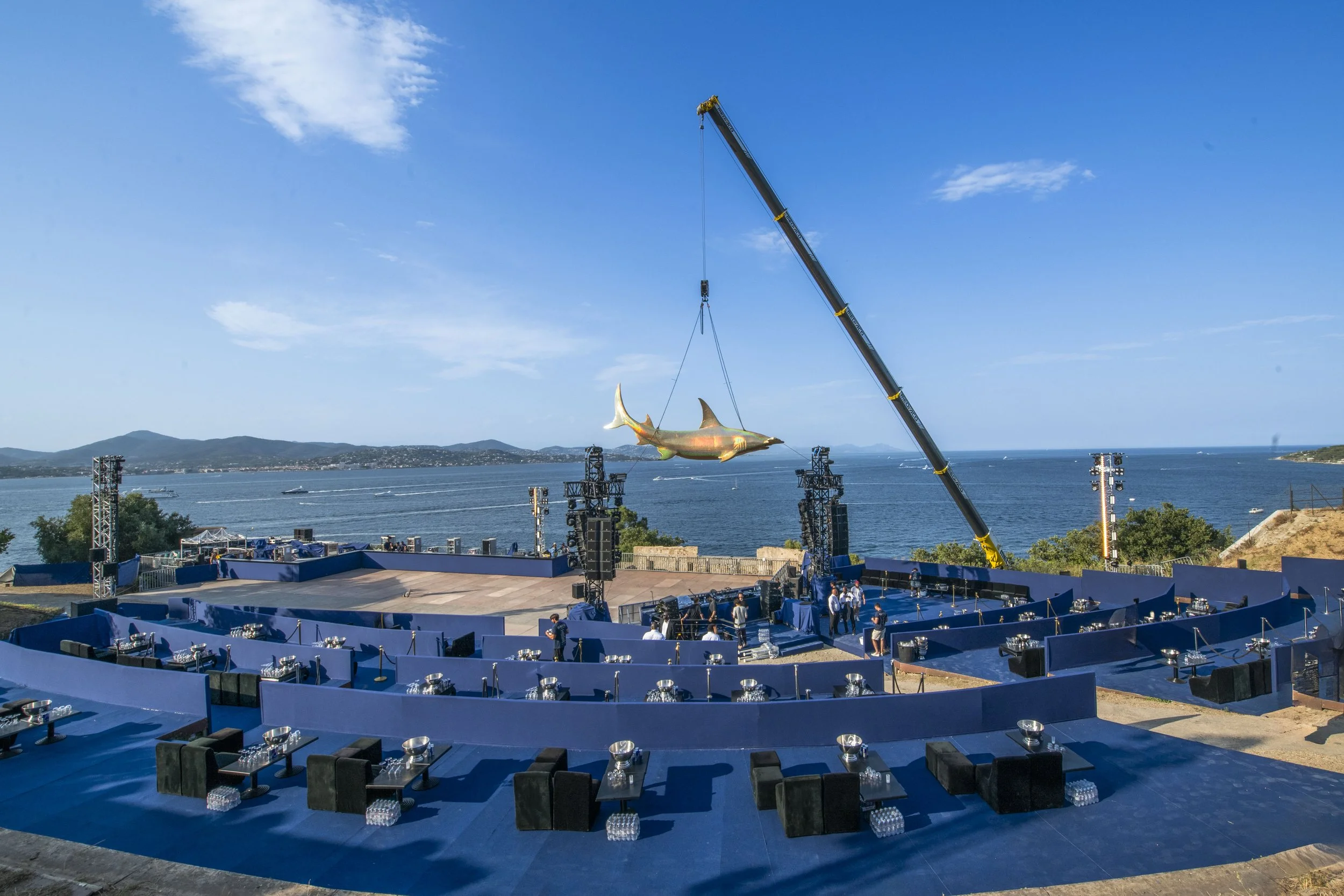 A large outdoor stage with blue flooring set up by the water, with tables and chairs arranged, a crane lifting a giant shark sculpture, and a scenic ocean view with mountains in the background.