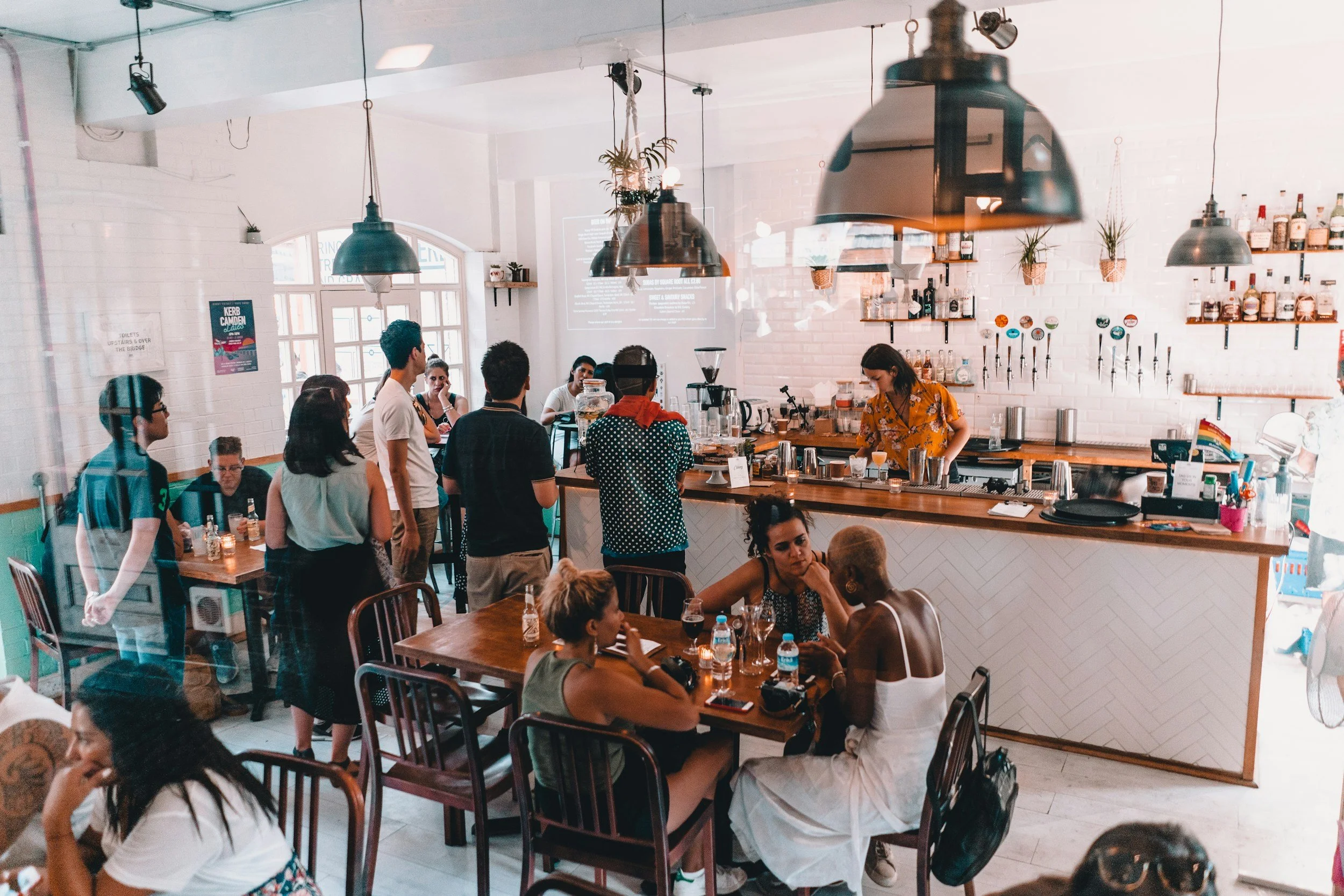 People ordering and socializing inside a modern coffee shop with white brick walls, hanging pendant lights, and baristas preparing drinks behind the counter.