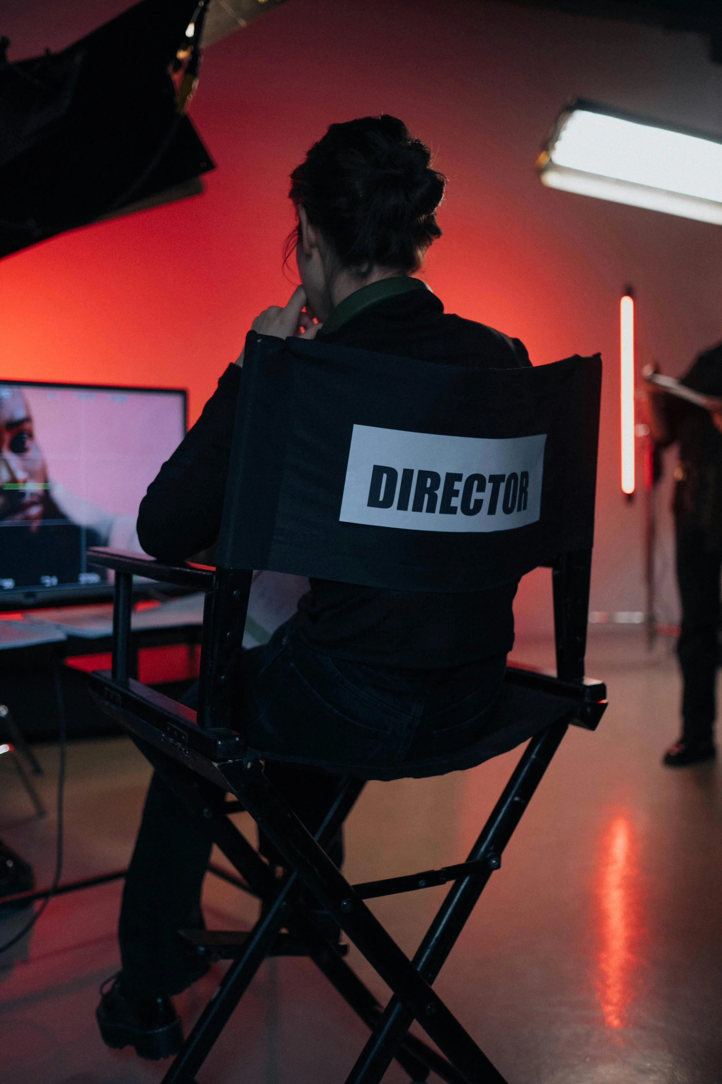 Film director sitting in a chair labeled 'Director' on a movie set with red lighting and equipment in the background.