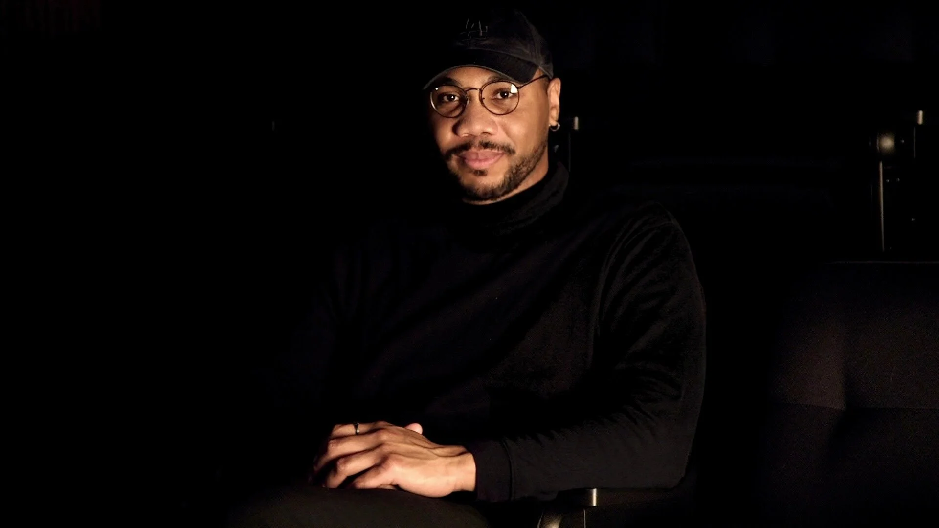 A man with glasses and a black cap sitting in a dark room, wearing a black turtleneck, with his hands clasped together.
