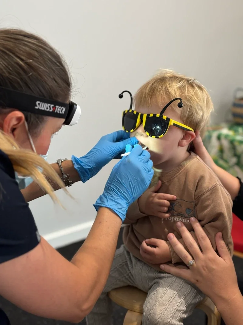 A healthcare professional examining a child wearing yellow sunglasses. The professional is using a light on their forehead and wearing blue gloves, checking the child's mouth, while another person gently supports the child.