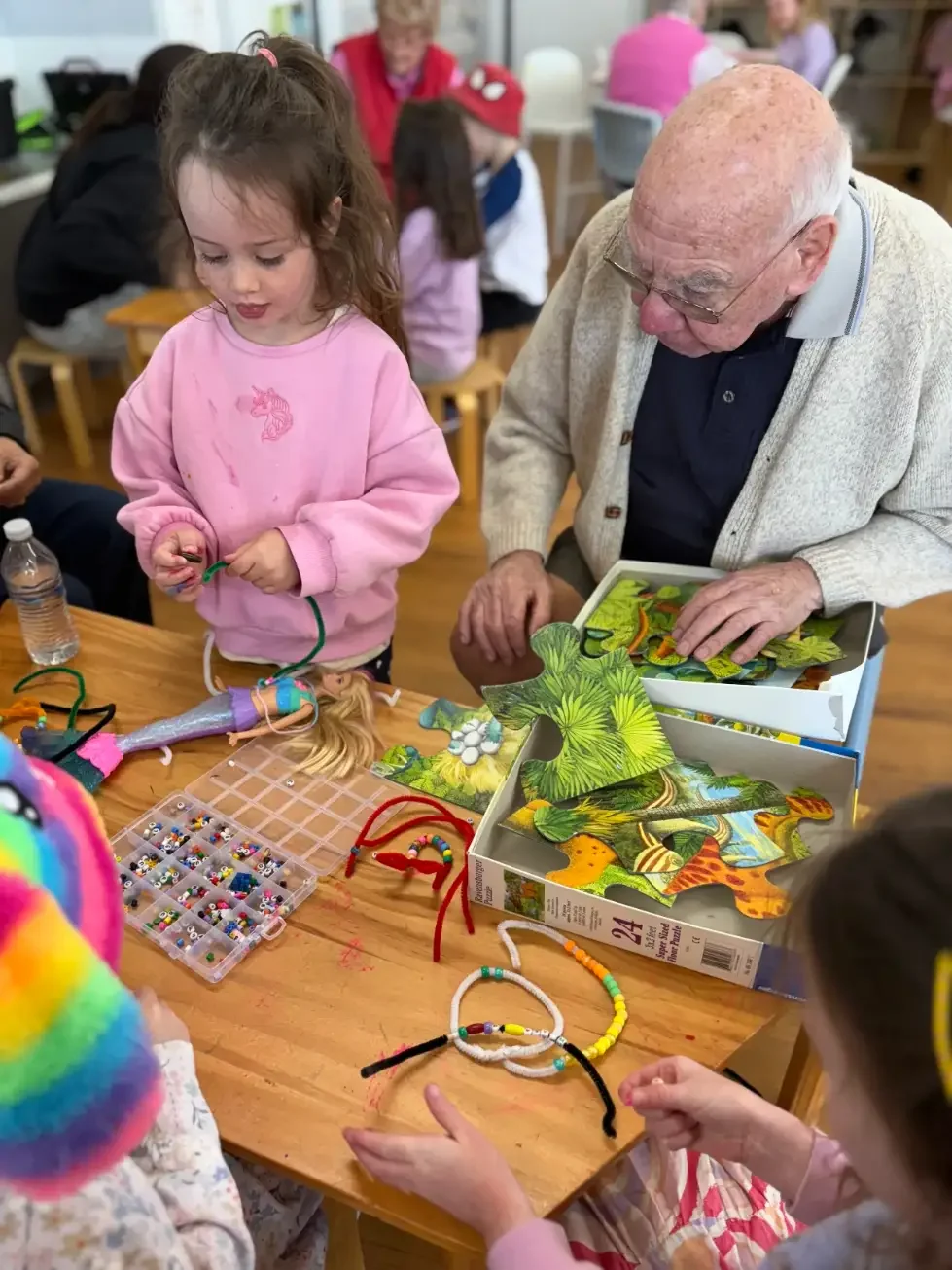 Children and an elderly man engaged in arts and crafts at a table, with jewelry-making supplies like beads, pipe cleaners, and jewelry pieces, in a classroom or craft room setting.