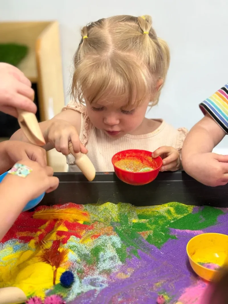 Children engaging in a sensory play activity with colorful sand and small objects.