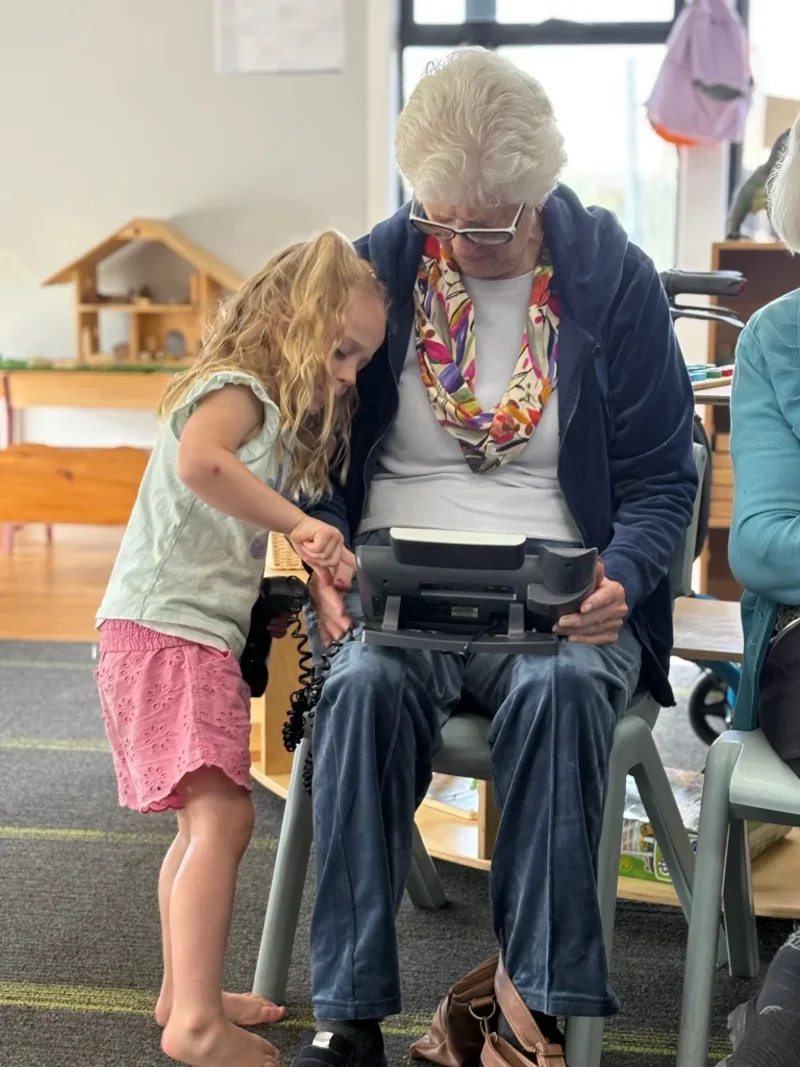 A child and elderly woman interacting with a handheld device, seated indoors.