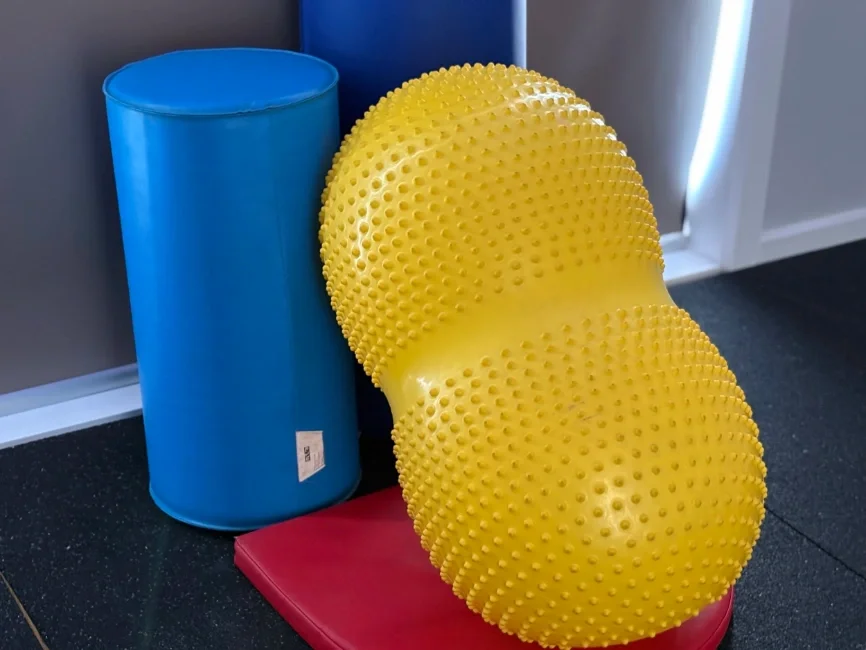 Gym equipment featuring a large blue cylinder, a textured yellow exercise ball, and a red mat on a gym floor.