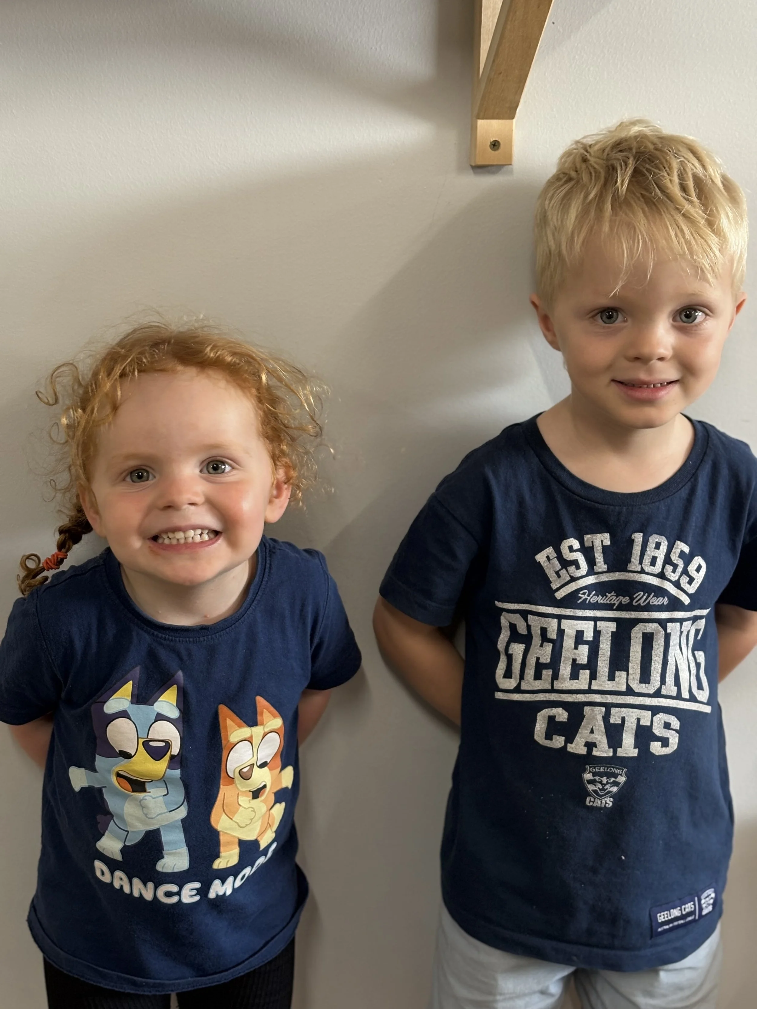Two young children standing indoors, smiling. The child on the left has curly red hair, wearing a navy blue shirt featuring animated characters. The child on the right has short blond hair, wearing a navy blue Geelong Cats T-shirt. A wooden wall bracket is visible above them.