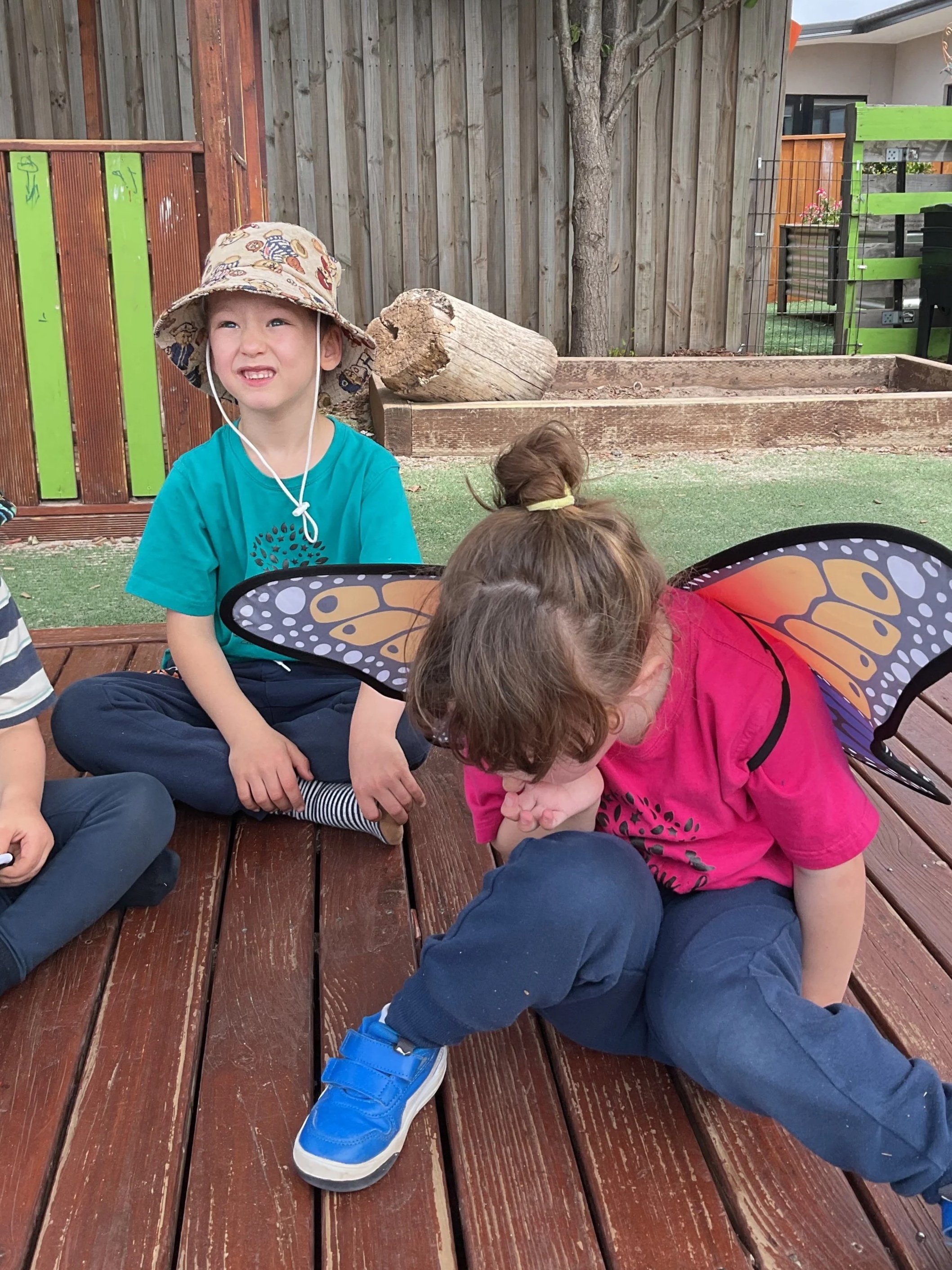 Two children sitting on a wooden deck, one with butterfly wings, the other wearing a sun hat and sitting cross-legged, with a backyard wooden fence and garden in the background.
