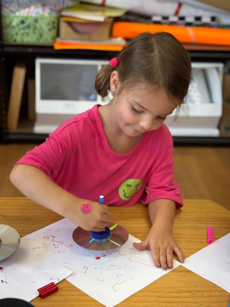 Young girl drawing with a CD spinner on paper at a table, wearing a pink shirt.
