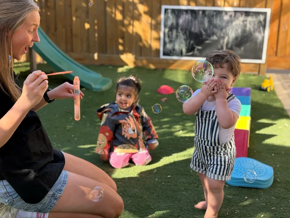 A woman blowing bubbles in a backyard with two children watching and playing.