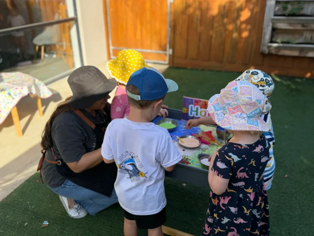 Group of children with a caregiver playing at a sensory table outside, wearing colorful hats and clothing.