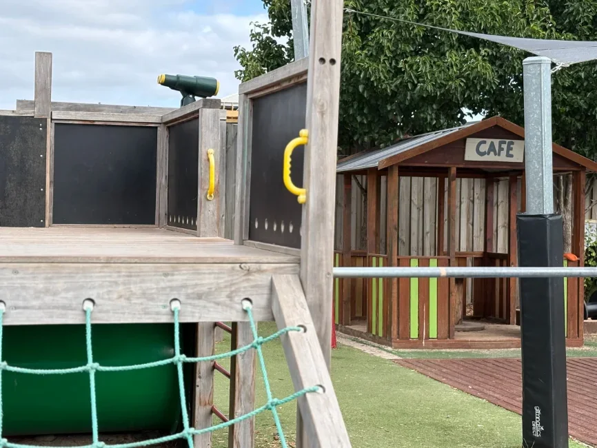 Playground with wooden structure, green slide, rope net, telescope, and a small play cafe in the background.