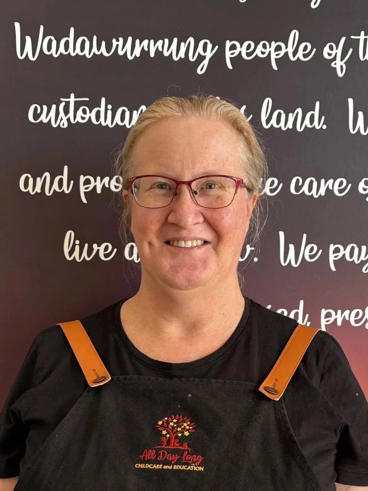 A woman with glasses smiling, wearing a black apron with a colorful tree logo and text, standing in front of a wall with white cursive writing about Wadahurrung people and land.
