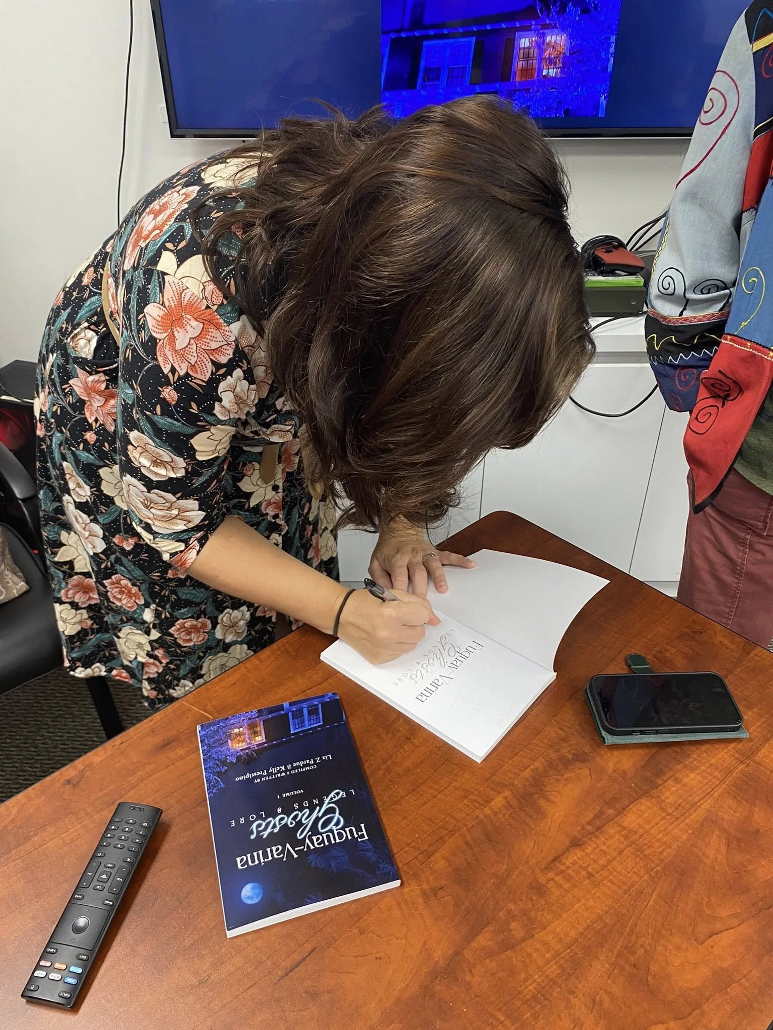 Person bending over a table, signing a book titled "Luxury-Vanity and the Gift of Perception". The table holds a remote control, a phone, and a booklet titled "Luxury-Vanity". Behind, a person in colorful clothing and a black shirt stand. In the back