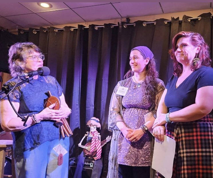 Three women standing on stage in front of a black curtain, engaging in a conversation. One woman is holding a wooden instrument and speaking into a microphone, while the other two women listen and smile. There is a small puppet or figurine in the bac