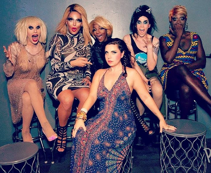 Six women dressed in colorful and fashionable outfits, sitting and posing together in a room with gray walls and black chairs.
