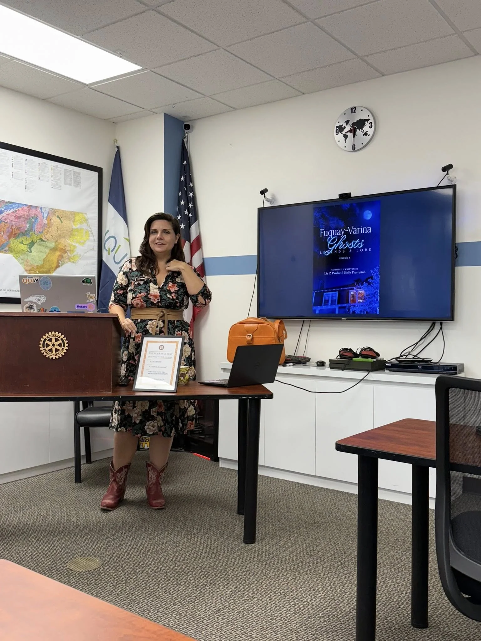 A woman wearing a floral dress and cowboy boots standing in a conference room with a computer, framed certificate, flags, and a large screen displaying the title 'Fuqay-Varina Ghosts' behind her.