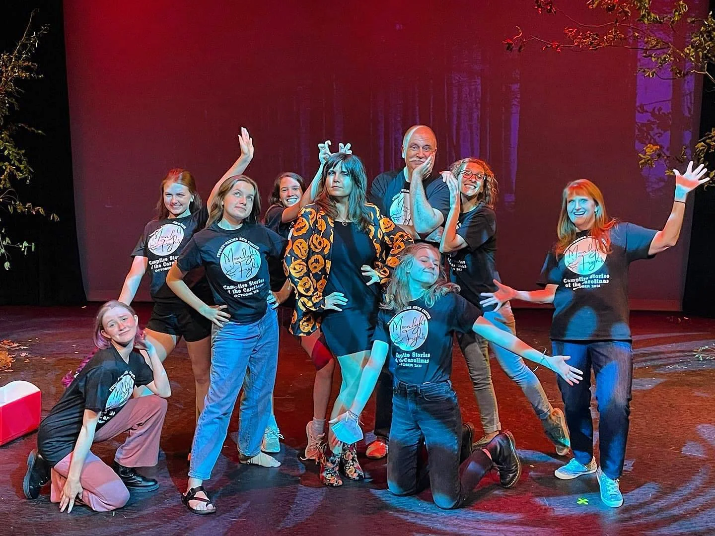 A group of people on stage, some wearing matching black t-shirts with a logo, posing with playful and happy expressions. The background features a forest scene projected on the screen.