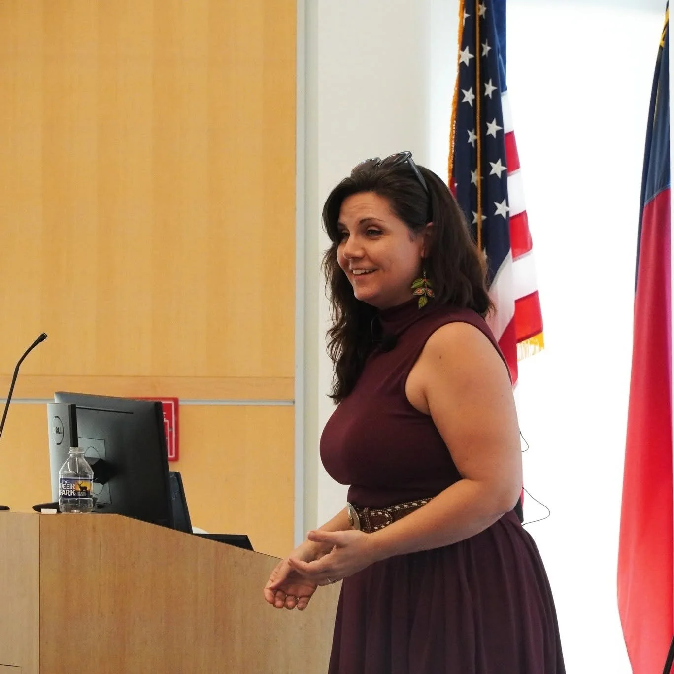 A woman with long dark hair in a sleeveless maroon dress, wearing colorful leaf-shaped earrings, standing and smiling in front of a podium in a conference room with American and state flags in the background.