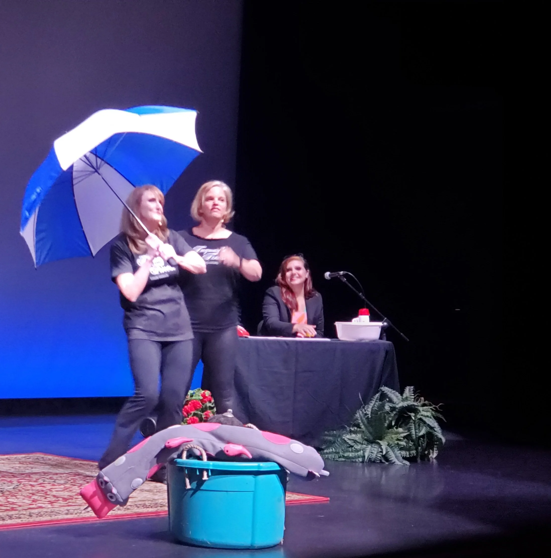 Two women on stage; one holding a blue and white umbrella, and another sitting at a table with a microphone. A third woman is sitting at the table and smiling. Items on stage include a carpet, a stuffed animal, a small blue cooler, and plants.