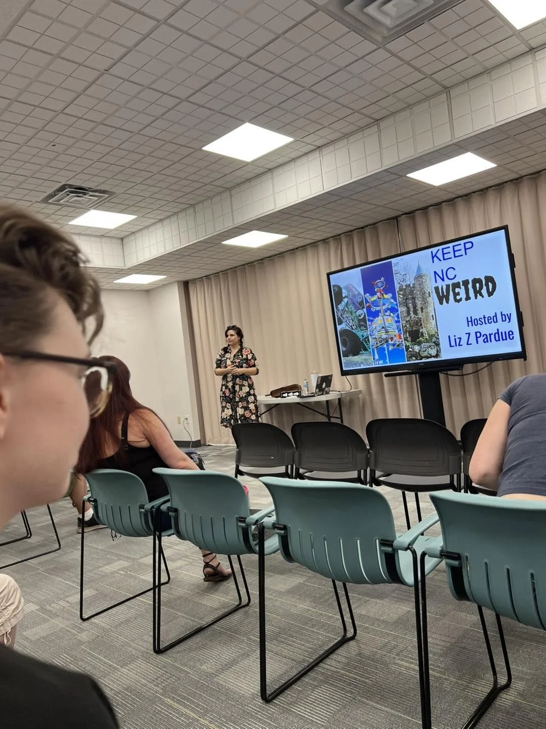 A woman stands at the front of a room giving a presentation. The screen next to her displays the words "KEEP NC WEIRD, Hosted by Liz Z Pardue." The audience seated faces the speaker, some with their backs to the camera.