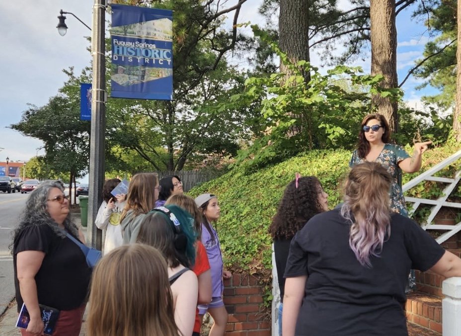 A woman with sunglasses talks to a group of people outside near a brick staircase, with a sign for Fuquay Springs Historic District hanging overhead.