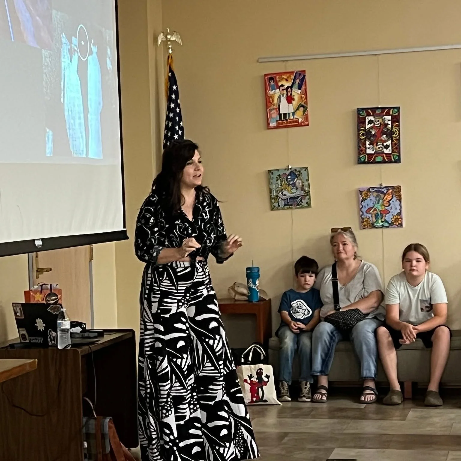 A woman in a black and white patterned dress is speaking in front of a group of children and an adult woman sitting on a bench, with colorful artwork hanging on the beige wall and an American flag in the background.
