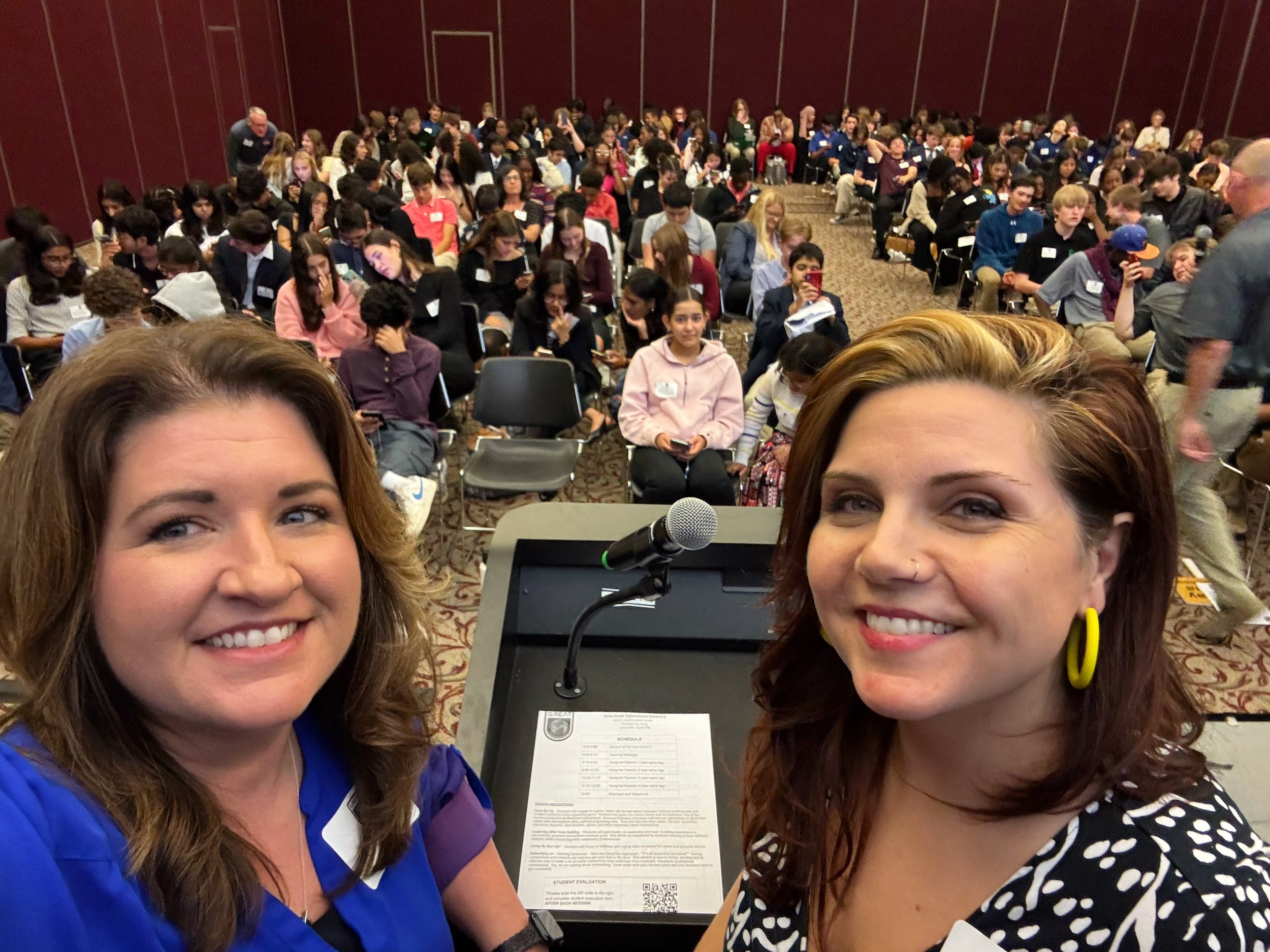 Two women taking a selfie in front of a microphone at a conference with many attendees seated behind them.