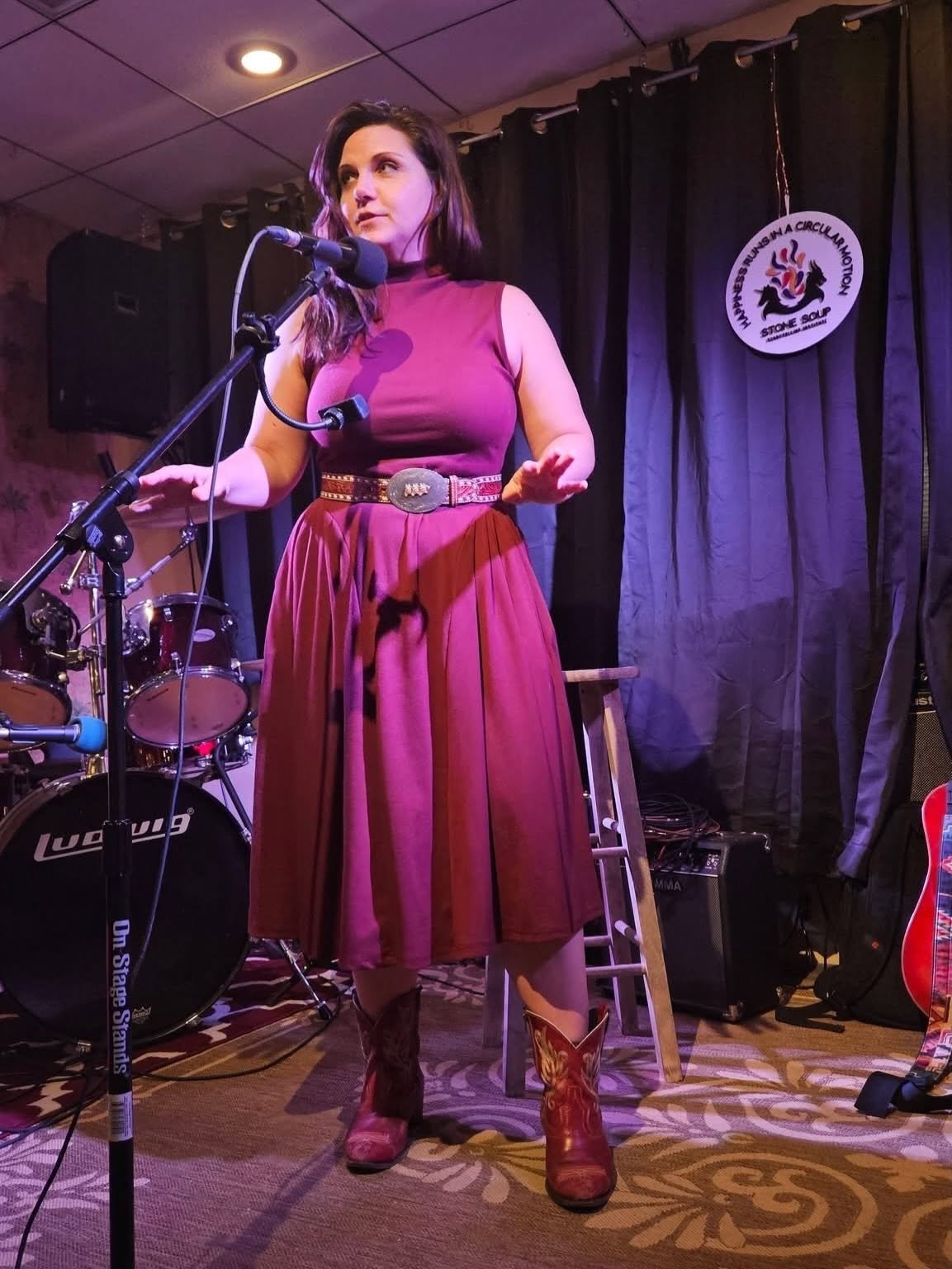 A woman speaking or singing into a microphone on stage, dressed in a sleeveless maroon top, long pleated maroon skirt, cowboy boots, and a wide belt, with a drum set and stage curtain in the background.