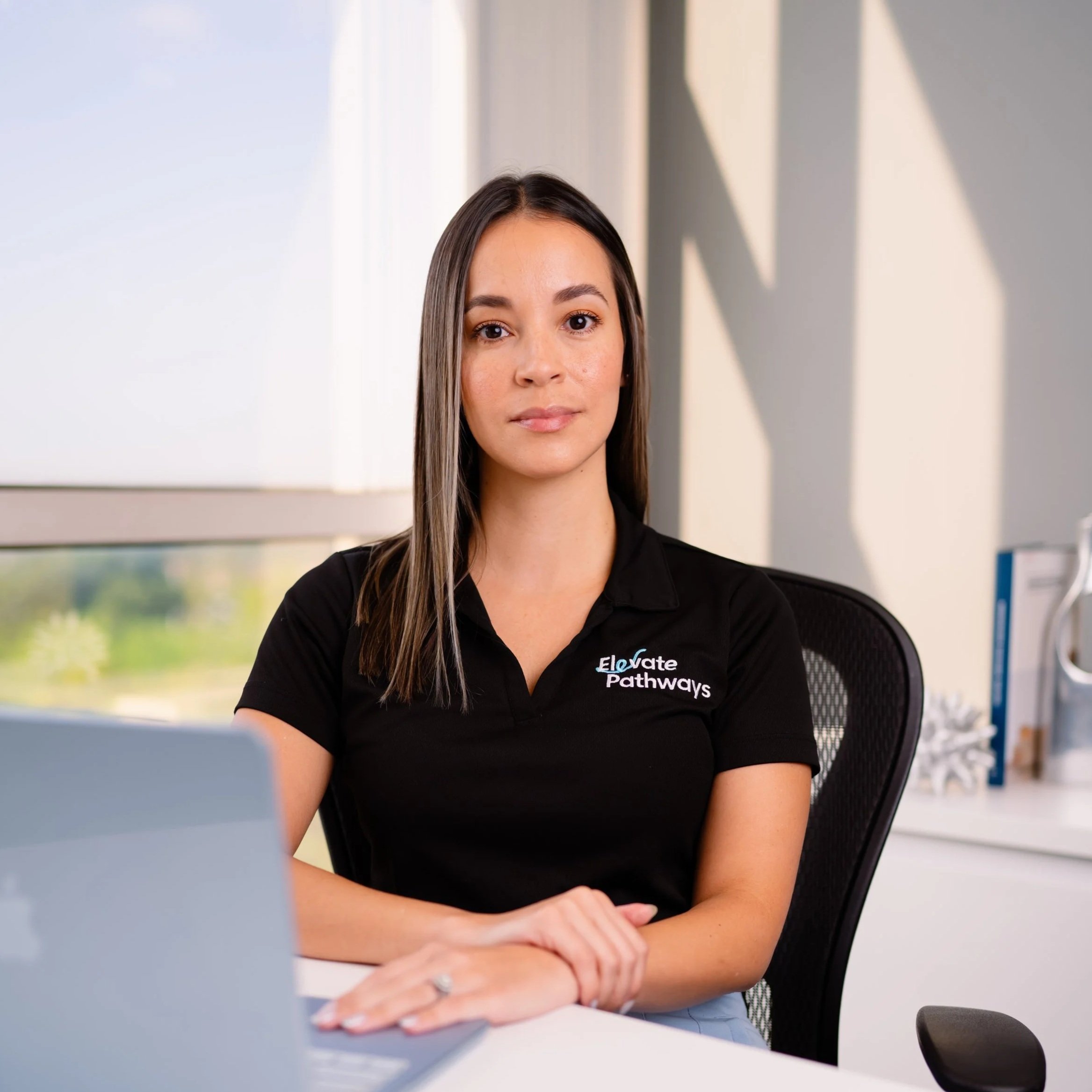 A woman with straight brown hair and light skin, wearing a black zip-up top, sitting against a light-colored wall.