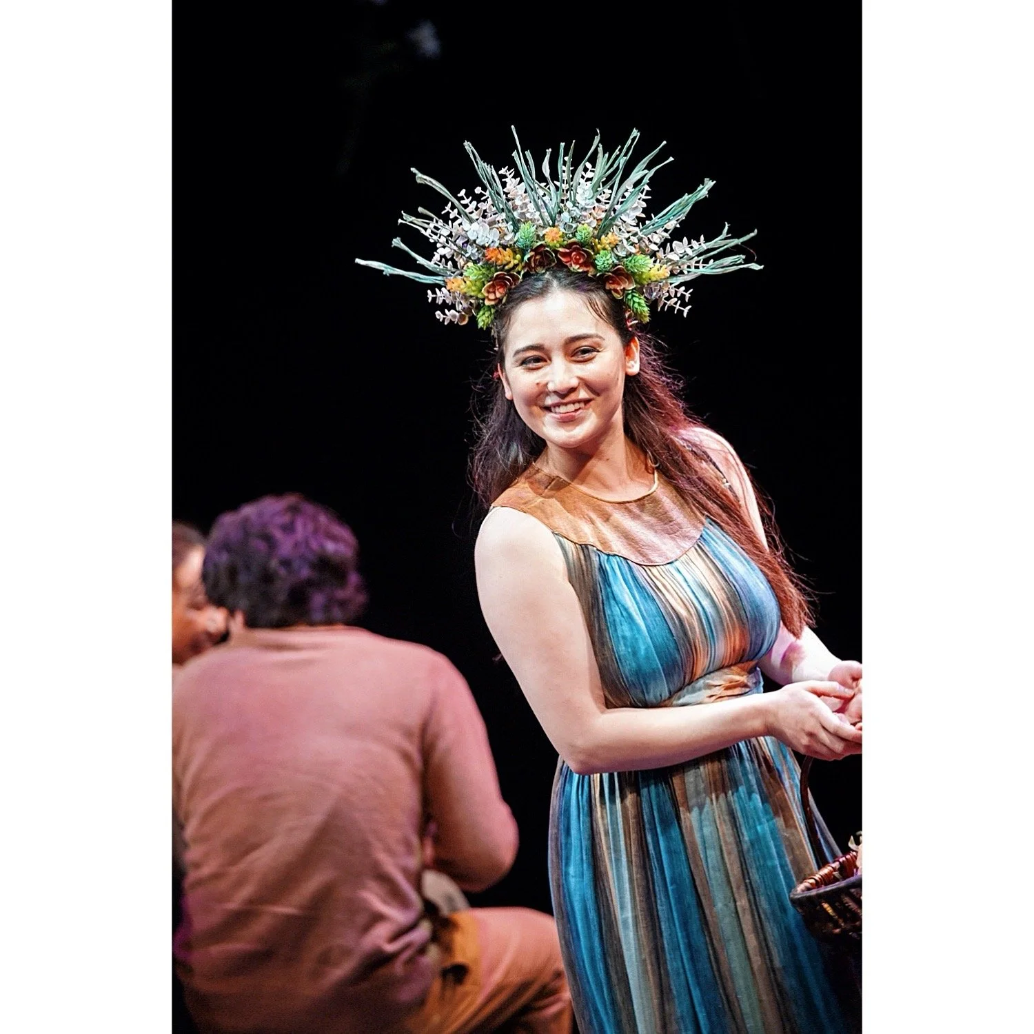 A woman wearing a floral headdress and a colorful, striped dress smiling on stage.