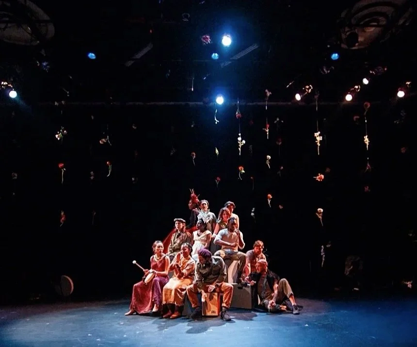A group of actors on stage during a theatrical performance, with hanging props above them and minimal lighting.