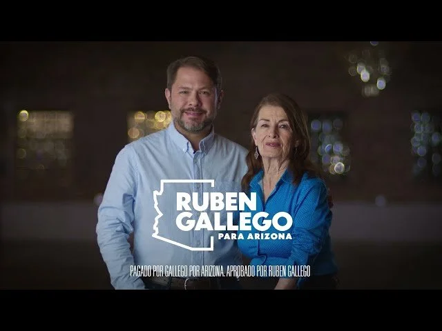 A man and woman standing together outdoors at night, with a blurred background of lights. The woman is wearing a blue shirt and the man is wearing a light blue shirt. There is a campaign sign with the text 'RUBEN GALLEGO PARA ARIZONA' and additional 
