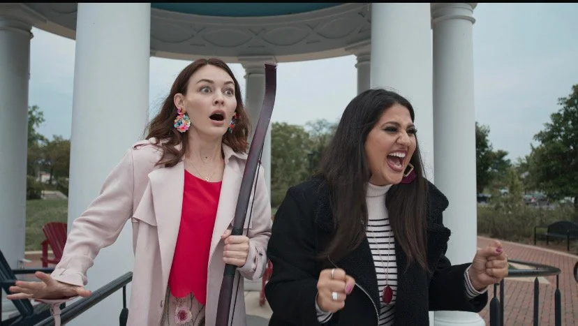 Two women are outdoors, appearing upset or angry, with one holding an umbrella. They are near a white gazebo with trees in the background.