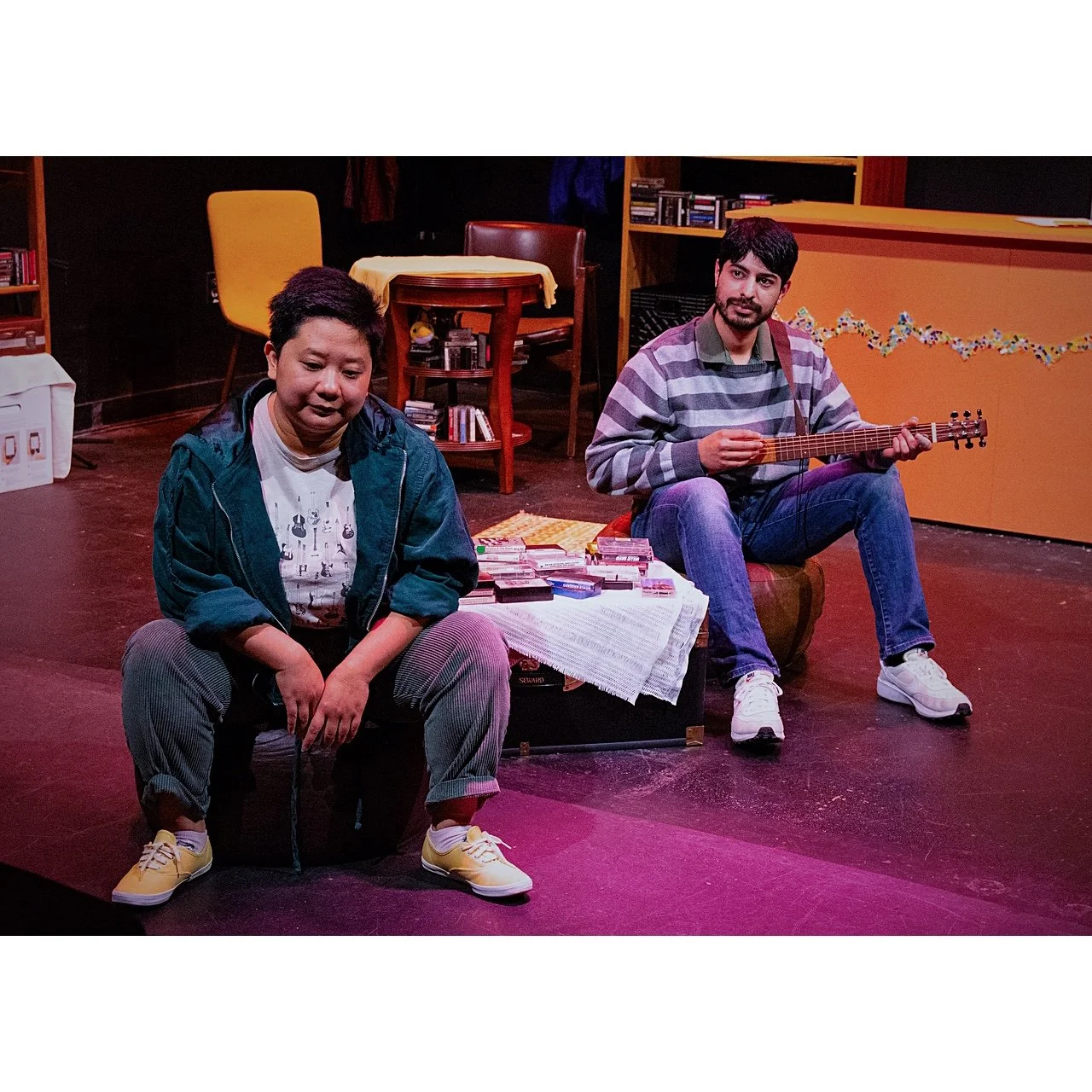 Two young men sitting on stage; one appears emotional, the other plays acoustic guitar. Stage has furniture and books.