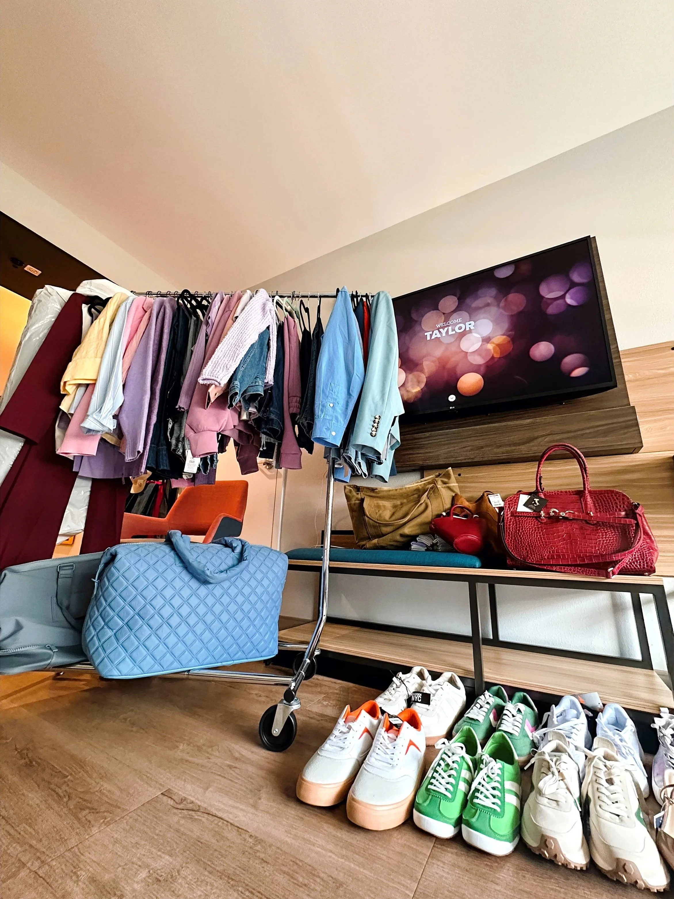 Clothing and accessories display, including a rack of various colored shirts, a table of handbags, and multiple pairs of sneakers on the floor under a TV with a welcome message for Taylor.