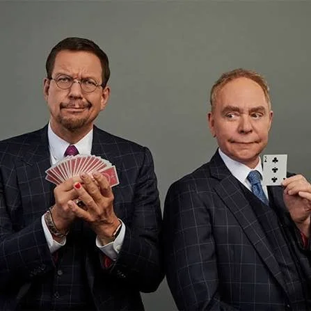 Two men in suits holding playing cards, one displaying a fan of cards and the other holding two aces, against a plain background.