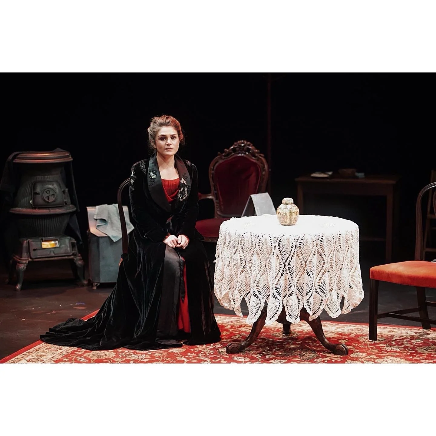 A woman dressed in period costume sitting on a chair next to a round table with a lace tablecloth and decorative jar. The setting appears to be a stage or theatrical performance space, with vintage furniture and a dark background.