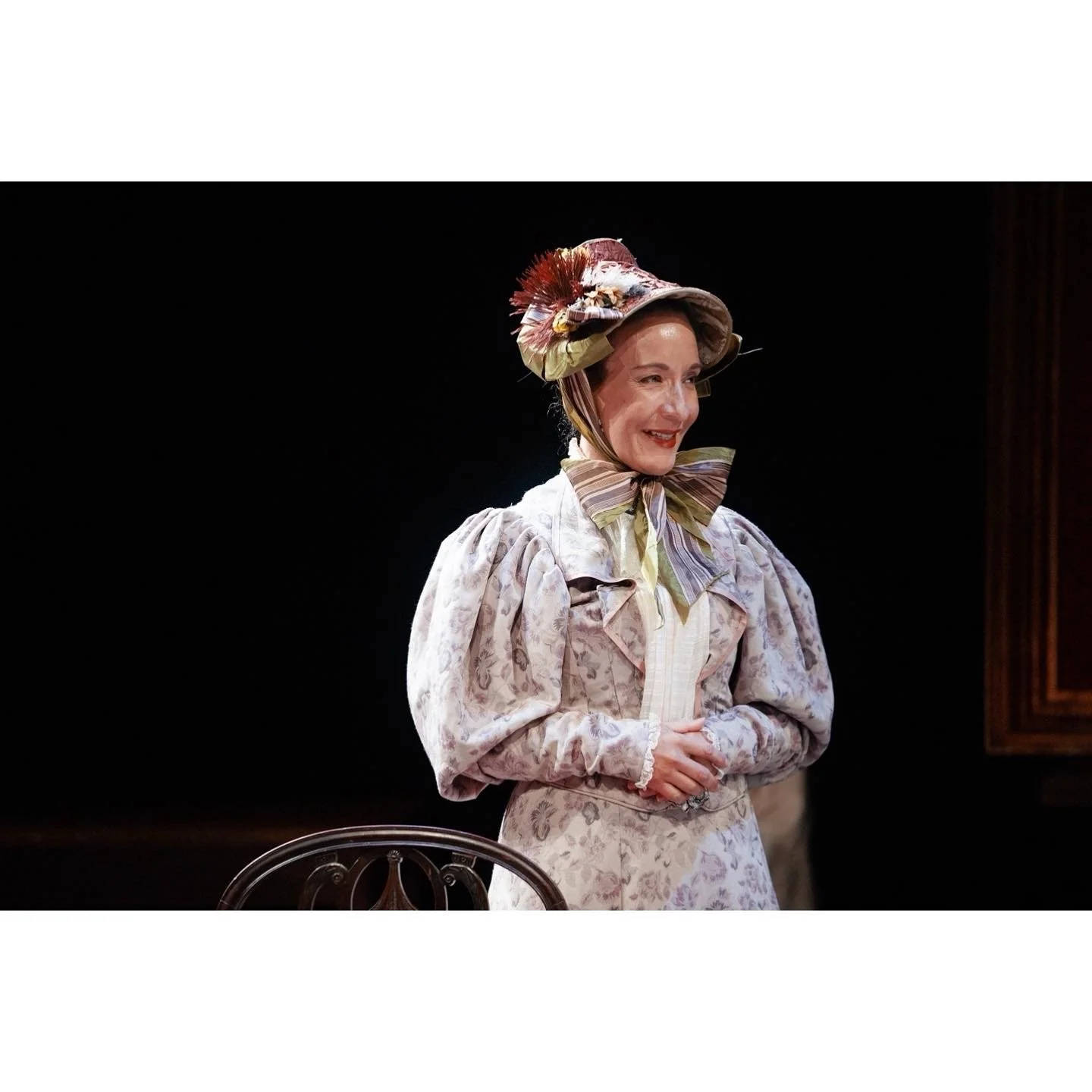A woman in a 19th-century costume with a floral print dress, puffed sleeves, a striped bow at her neck, and a large floral hat, smiling in a vintage setting.