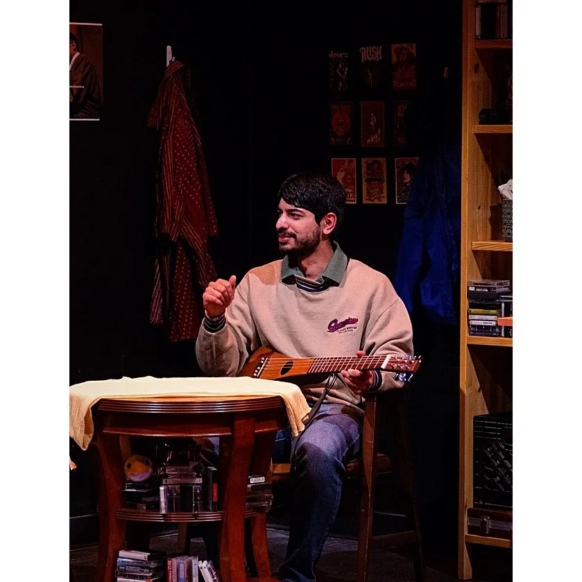 Young man playing acoustic guitar and smiling in a cozy room with bookshelves, posters, and jackets hanging on the wall.